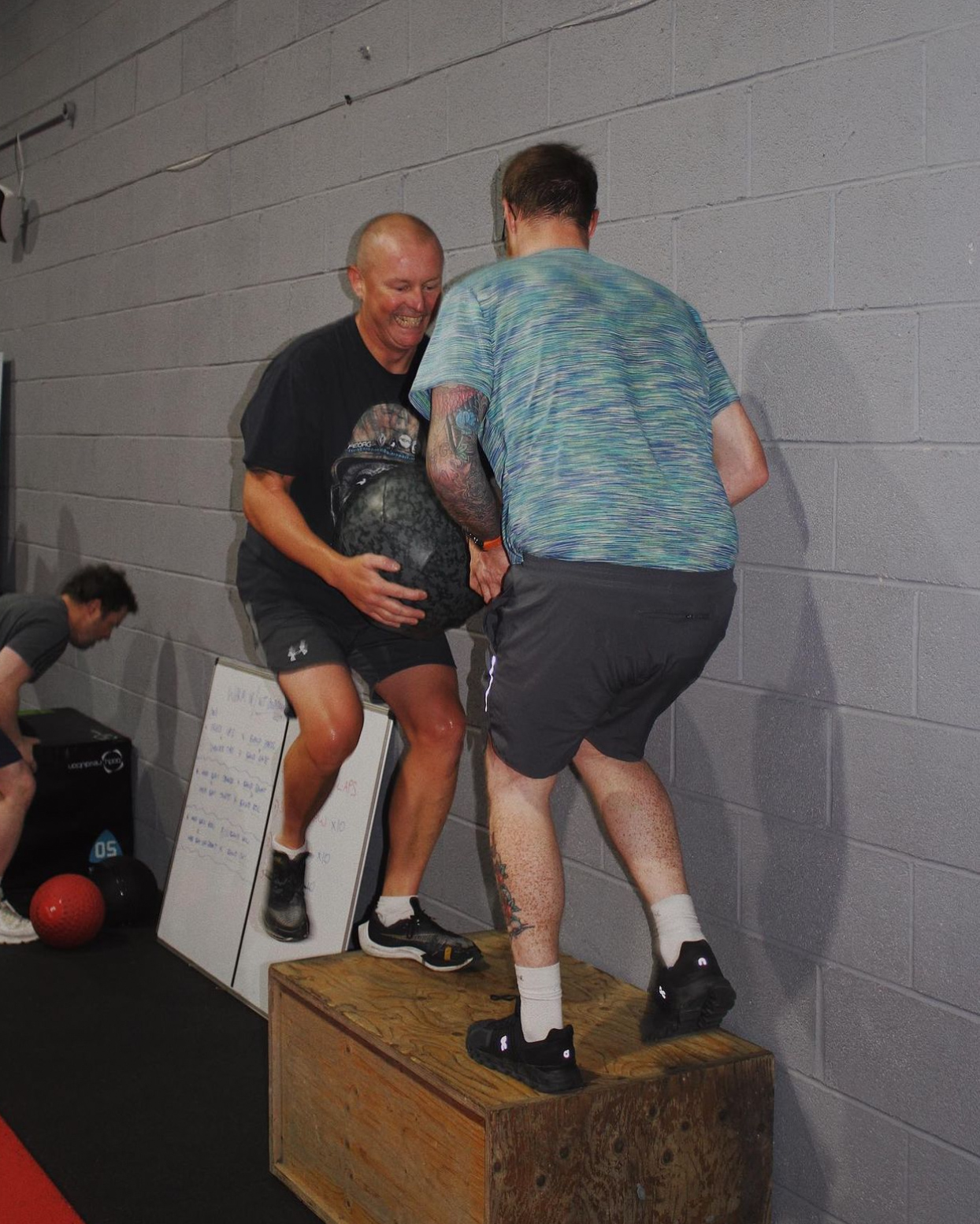 Two men in a conditioning class, smiling and cheering each other on while performing step-ups. This image represents the fun and social aspects of group fitness classes.